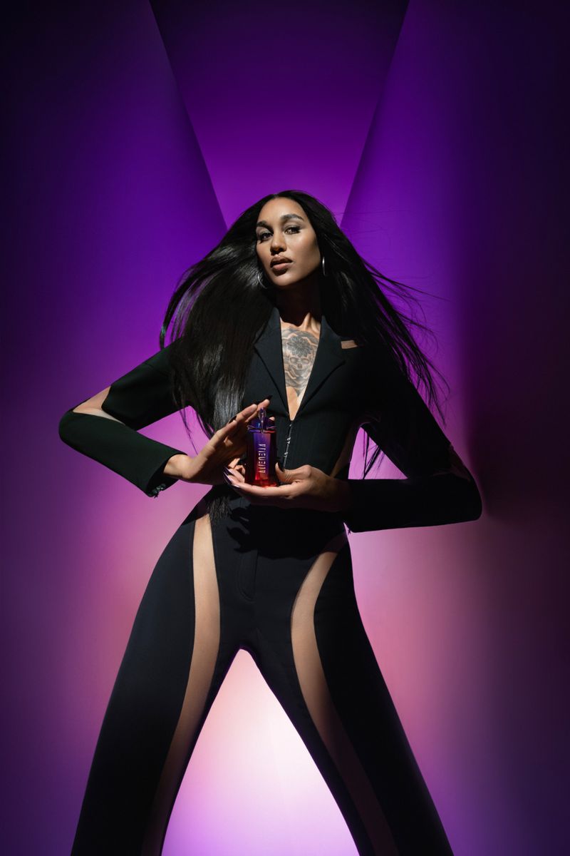 Elegant adult female posing with a perfume bottle against a purple background in a studio, wearing a chic black pantsuit.