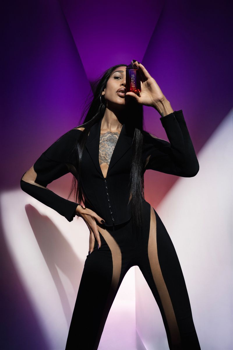 Adult woman in a black cutout suit, holding a red bottle in a photo studio setting with a purple and white geometric background.