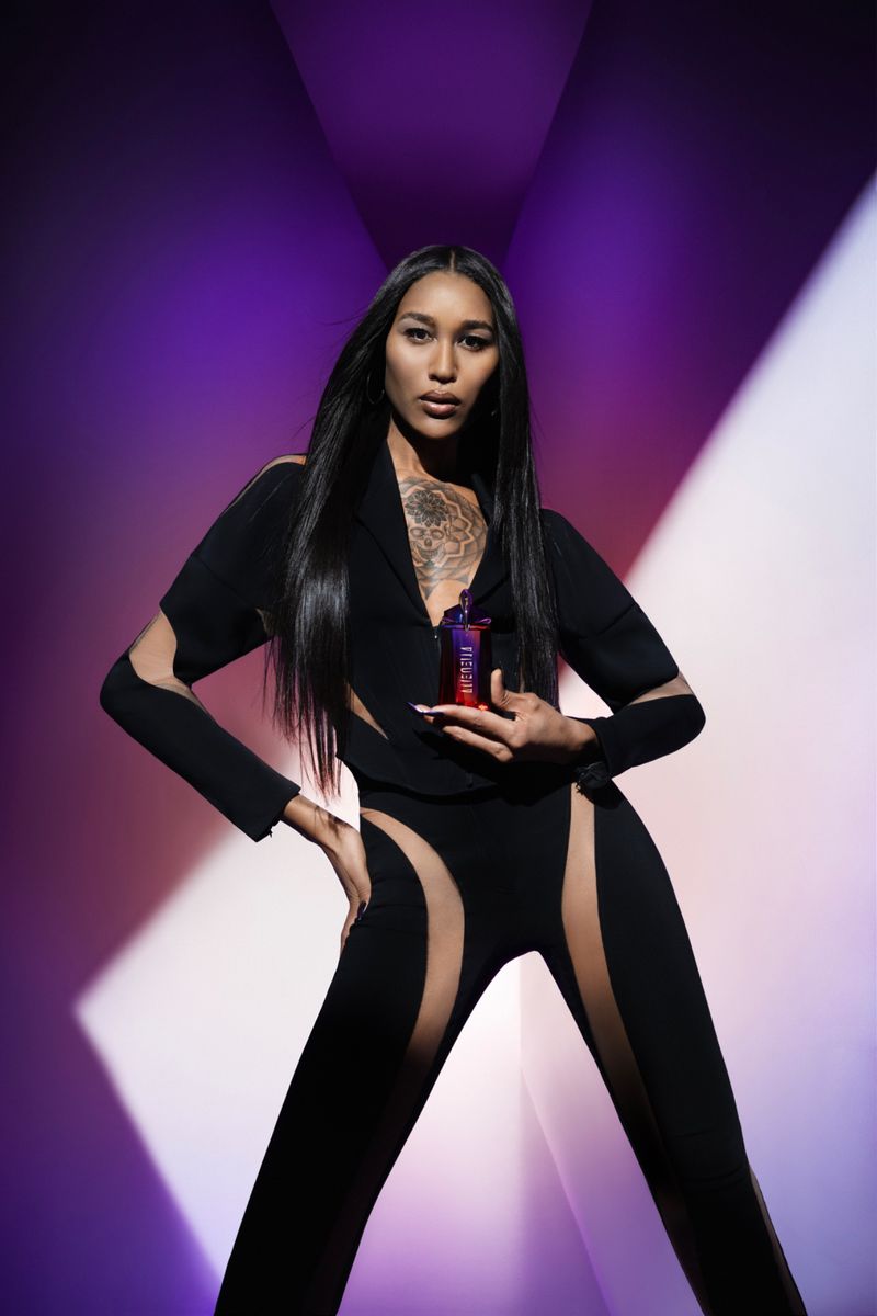 Woman in black jumpsuit holding perfume bottle with purple and orange gradient in a studio with purple and white lighting.