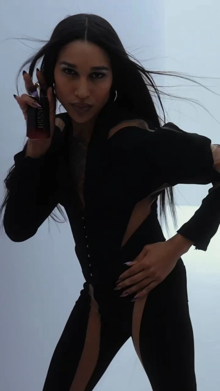 Model Gia Bab holding a Mugler perfume bottle wearing a black outfit against a plain white background