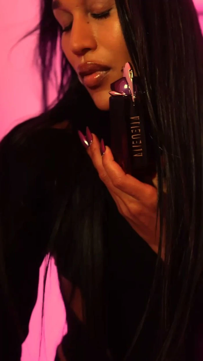 Model Gia Bab holding Mugler Alien Hypersense perfume bottle against a vibrant pink backdrop for Mugler campaign.