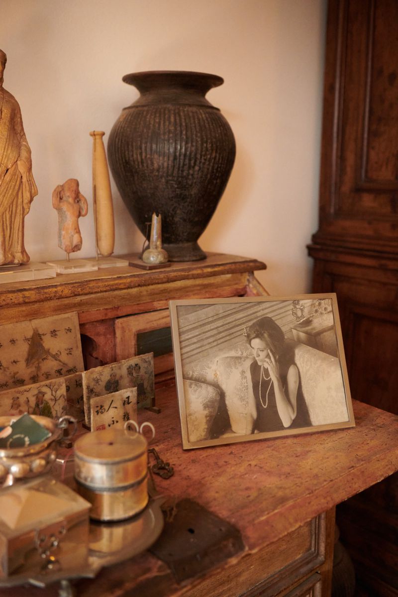 Indoor still life showcases a framed photo of a woman, antique vase, and small figurines on a wooden desk, with a warm, vintage aesthetic.