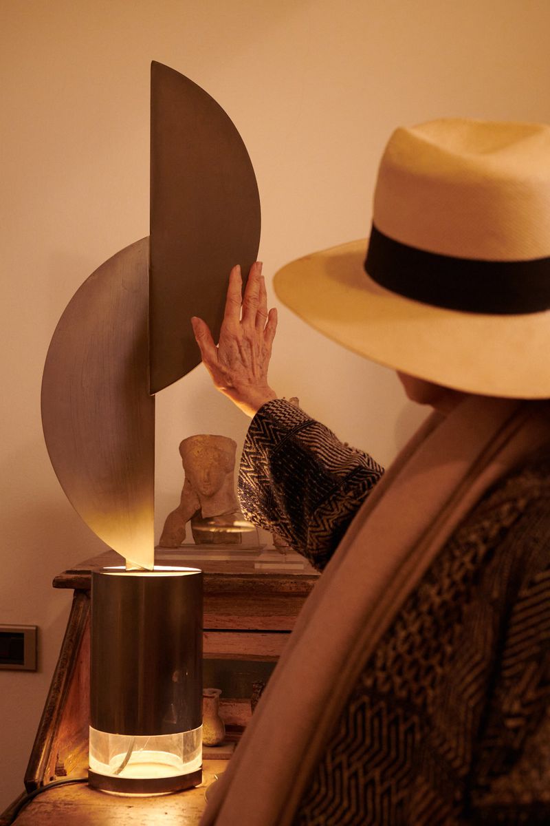Woman in straw hat adjusts modern lamp atop wooden desk with bust sculpture in warm-toned interior, creating soft, artistic lighting.