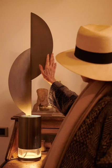 Woman in straw hat adjusts modern lamp atop wooden desk with bust sculpture in warm-toned interior, creating soft, artistic lighting.