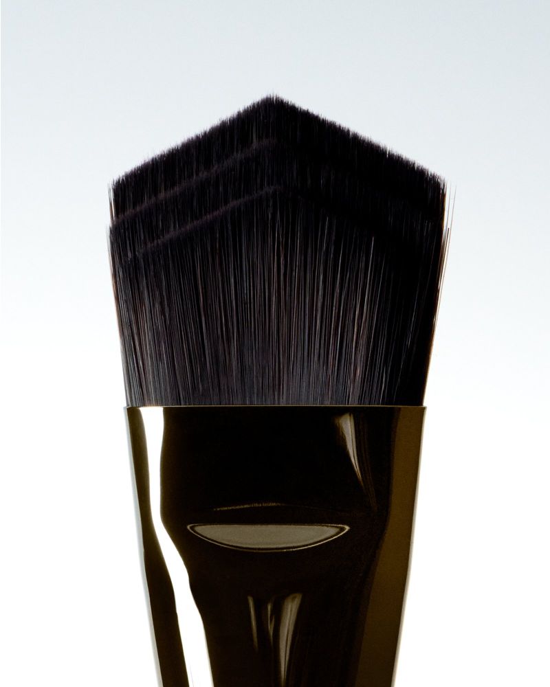Close-up studio shot of a makeup brush with black bristles and a shiny gold handle against a clean white background.