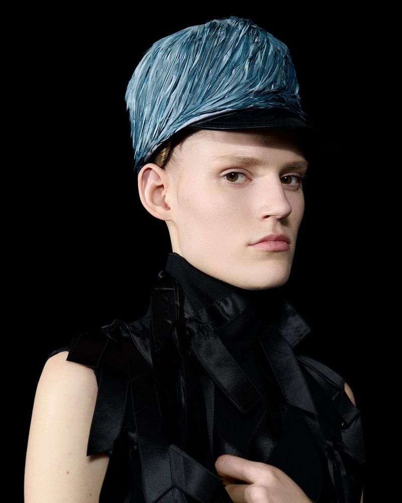 Headshot of a fair-skinned person wearing a textured blue hat and black sleeveless top against a dark background in a studio setting.