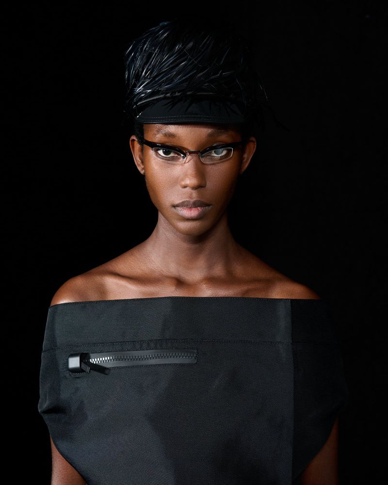 Fashion portrait of a Black adult female model wearing a feathered hat, glasses, and black off-the-shoulder top against a dark background.