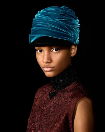 Portrait of a woman with dark skin wearing a teal head covering and burgundy floral dress against a solid black background.