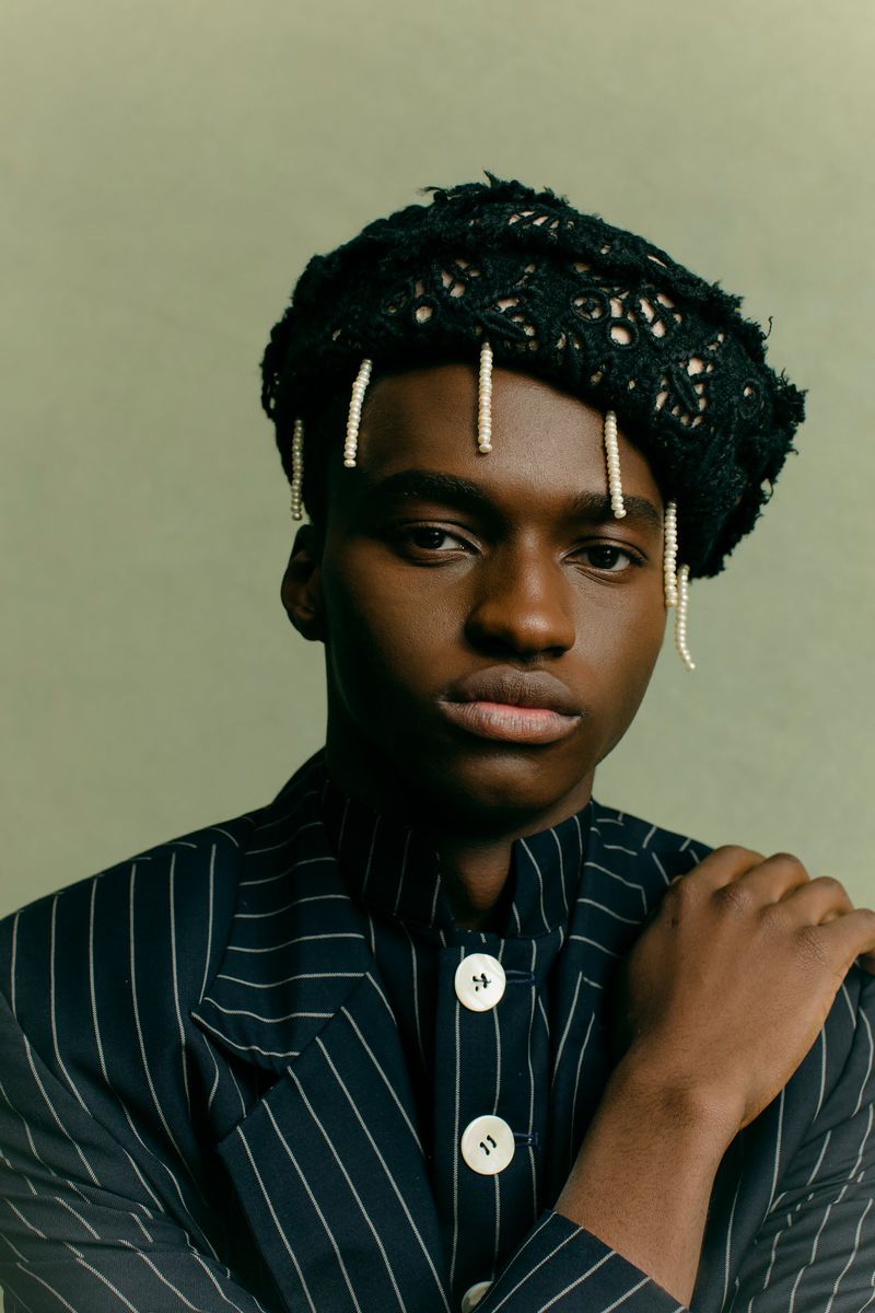 Portrait of a black male model in pinstripe jacket and black knit hat, posing indoors against a solid green background.