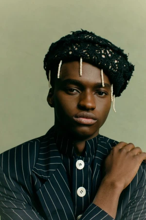 Portrait of a black male model in pinstripe jacket and black knit hat, posing indoors against a solid green background.
