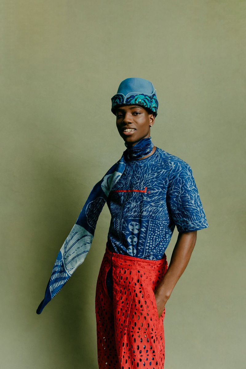 A fashionable Black man with a patterned blue shirt, hat, and scarf poses indoors against a muted green backdrop, wearing red lace pants.