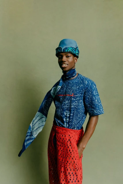 A fashionable Black man with a patterned blue shirt, hat, and scarf poses indoors against a muted green backdrop, wearing red lace pants.