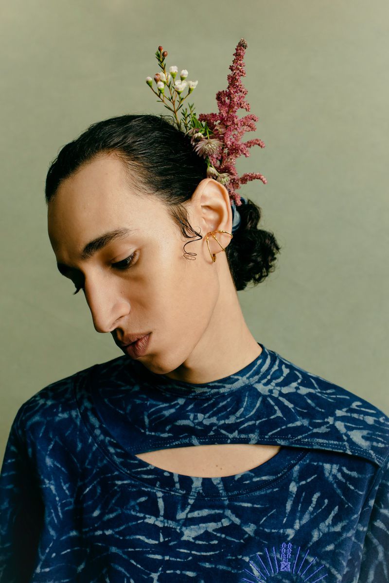 Portrait of a person with flowers in their hair, wearing a patterned blue shirt against a muted green background in soft studio lighting.