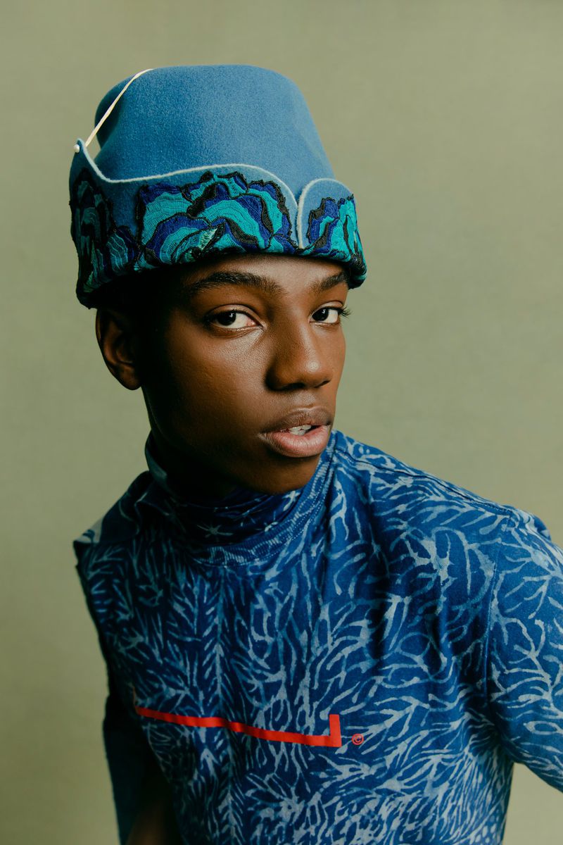 Fashion portrait of a Black person with a patterned shirt and hat against a light green background in a studio setting.