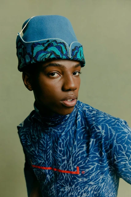 Fashion portrait of a Black person with a patterned shirt and hat against a light green background in a studio setting.