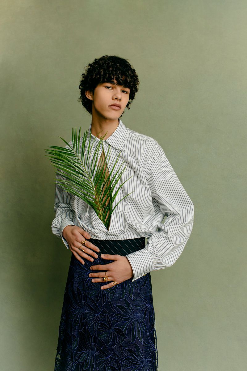 Androgynous fashion portrait with person in striped shirt holding palm leaf, set against a solid light green background, studio lighting.