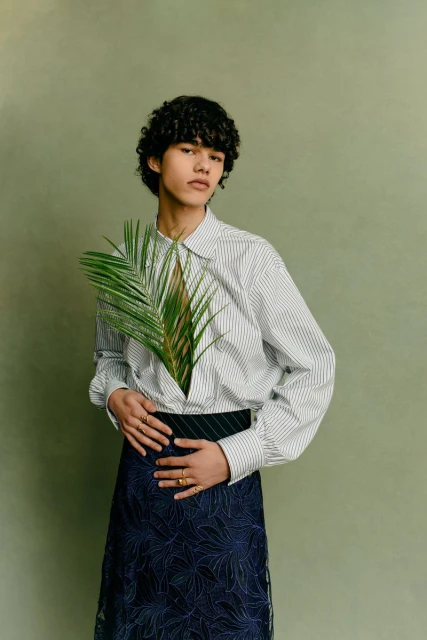 Androgynous fashion portrait with person in striped shirt holding palm leaf, set against a solid light green background, studio lighting.