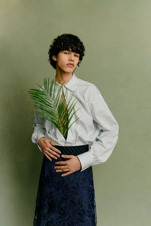 Androgynous fashion portrait with person in striped shirt holding palm leaf, set against a solid light green background, studio lighting.