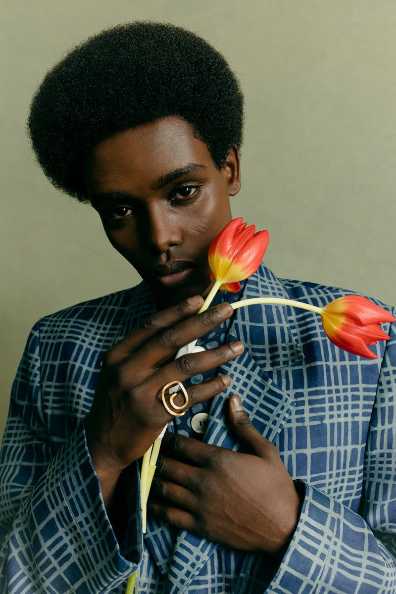 Close-up portrait of a Black male model in a blue checkered blazer holding two red tulips, wearing a gold ring, against a muted green backdrop.