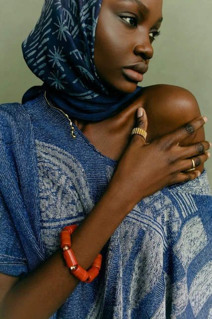 Close-up of a Black woman with a patterned headscarf, wearing a blue patterned top, orange beaded bracelet, and gold rings, against a green background.