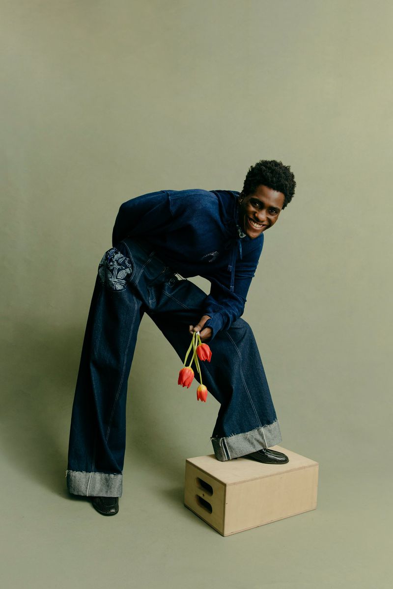 Smiling adult poses in denim outfit, standing on box holding red tulips against a muted green backdrop in a studio setting.