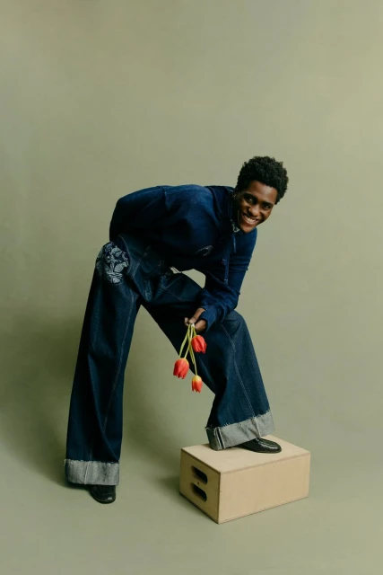 Smiling adult poses in denim outfit, standing on box holding red tulips against a muted green backdrop in a studio setting.