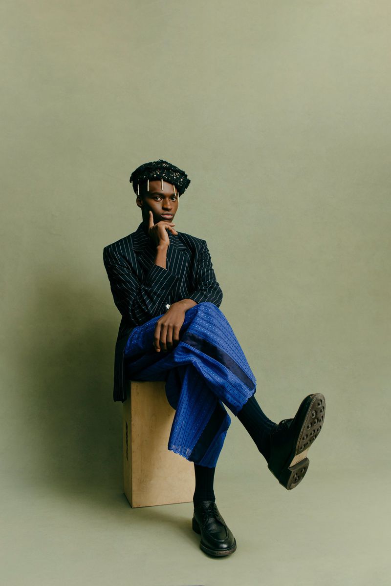 Adult male in pinstripe blazer and blue culottes sitting on a box in a photo studio against an olive-green backdrop.
