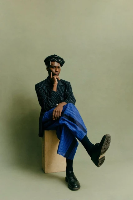 Adult male in pinstripe blazer and blue culottes sitting on a box in a photo studio against an olive-green backdrop.