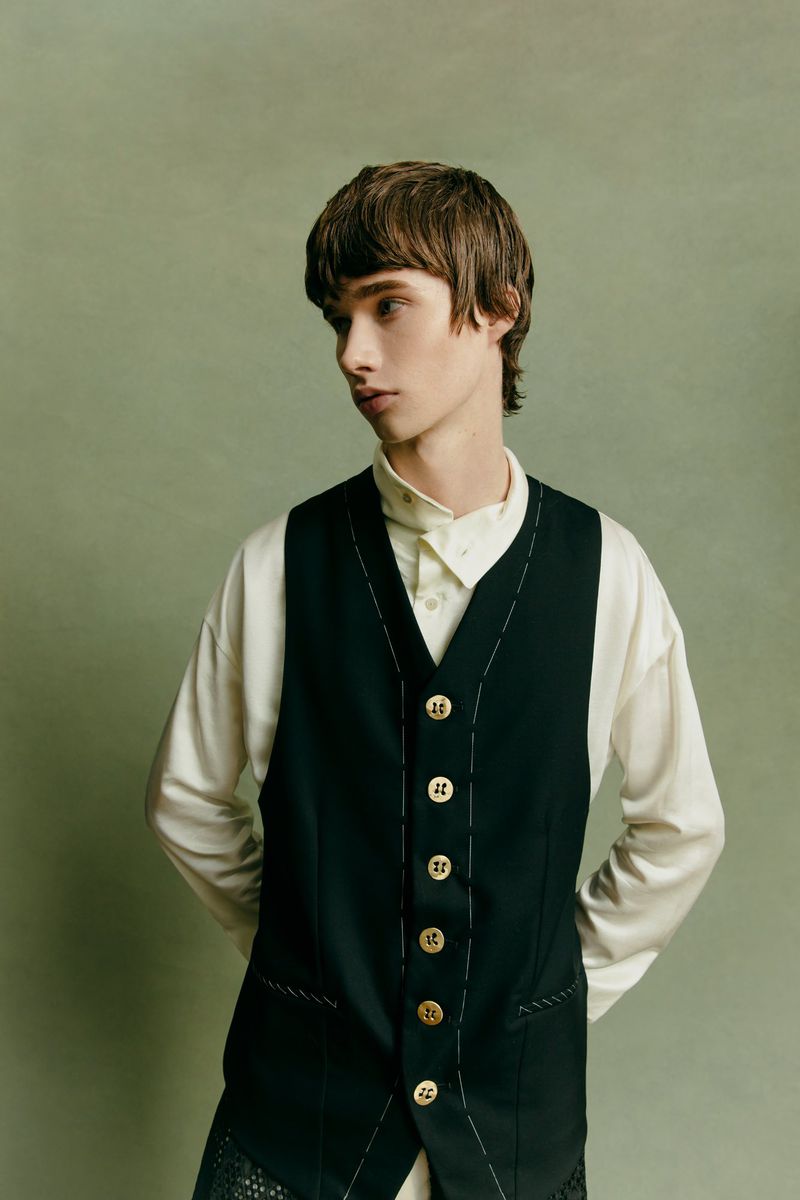 Studio shot of a young male model in a black vest with gold buttons and a white shirt, posing against a green background, looking away.