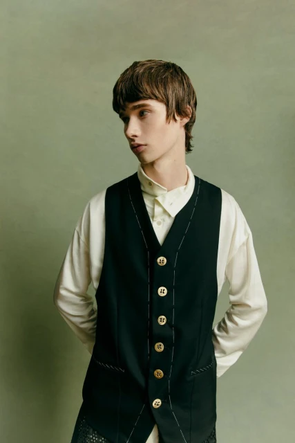 Studio shot of a young male model in a black vest with gold buttons and a white shirt, posing against a green background, looking away.