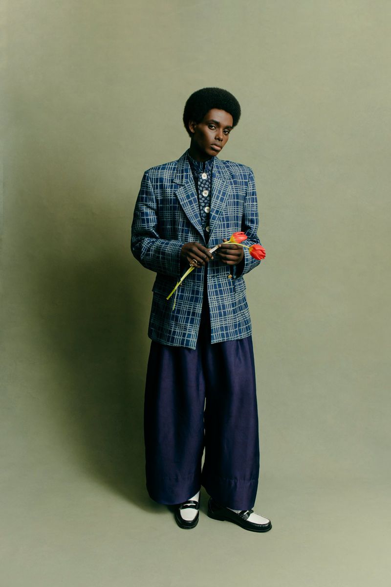 Young adult with an afro poses in a blue plaid blazer and wide-legged pants while holding red tulips against a green background.