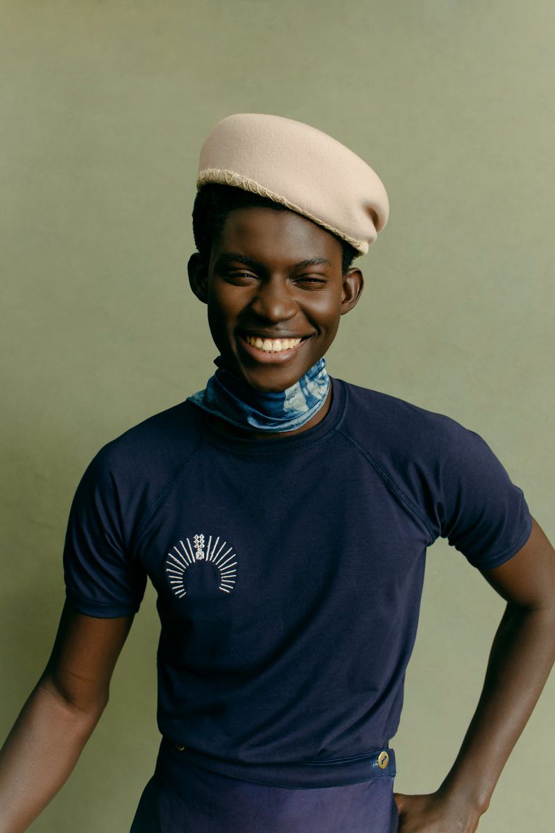 A smiling African-American adult male wearing a navy t-shirt, blue neck scarf, and beige beret poses indoors against an olive green wall.