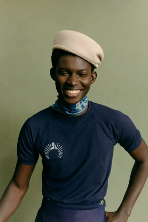 A smiling African-American adult male wearing a navy t-shirt, blue neck scarf, and beige beret poses indoors against an olive green wall.