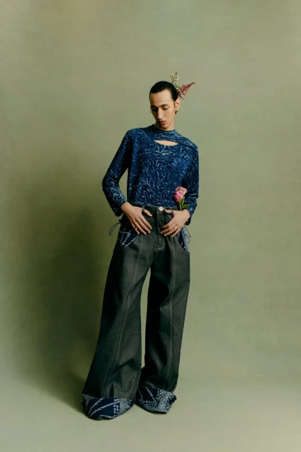 Fashionable person in a patterned blue top and grey wide-leg jeans strikes a pose in a studio with flowers and a muted green backdrop.