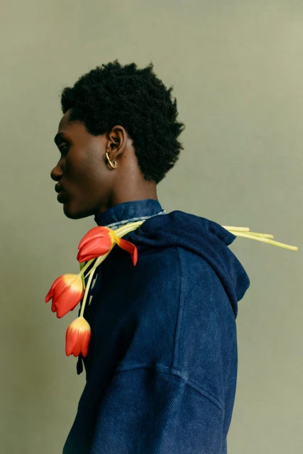 Profile shot of a Black male model in denim hoodie with red and yellow tulips draped over his shoulder in a fashion editorial.