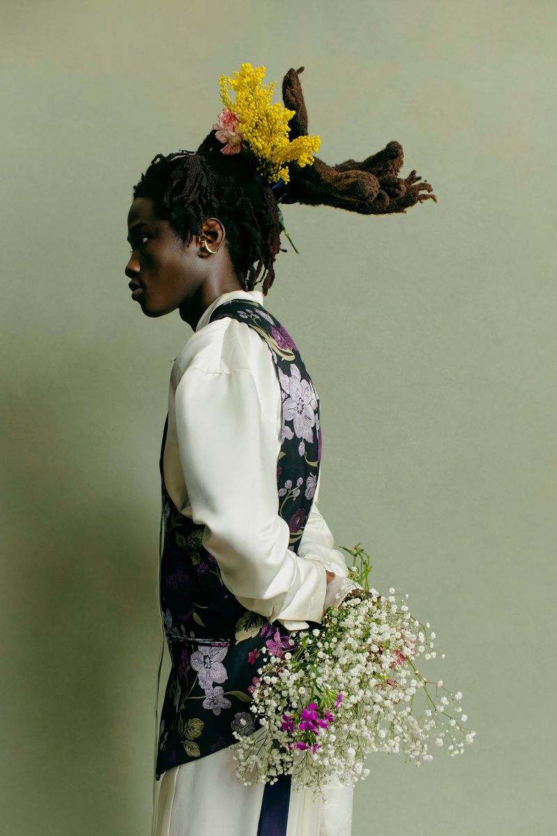 Profile shot of a Black adult male wearing floral vest and white clothing, holding a bouquet with flowers in his hair against green backdrop.