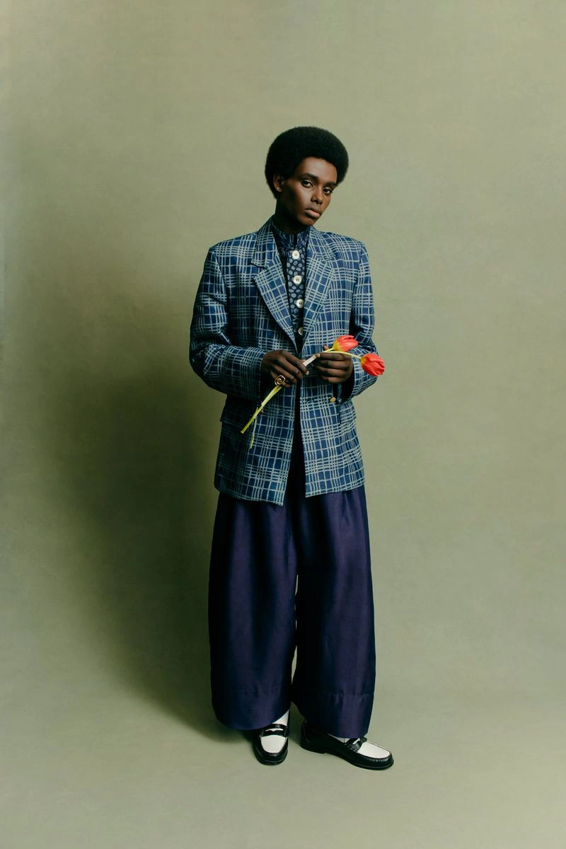 Model holding tulips wearing a patterned plaid blazer and wide trousers against a solid olive backdrop
