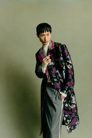 Model in a floral patterned coat and grey trousers posing against a muted green studio backdrop.