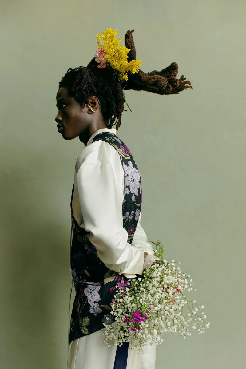 Profile view of a black man wearing a floral patterned vest and white shirt holding a bouquet of flowers against a green backdrop