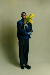 Model holding a bright yellow bouquet of flowers, wearing a blue patterned top and striped trousers against a muted green studio backdrop.