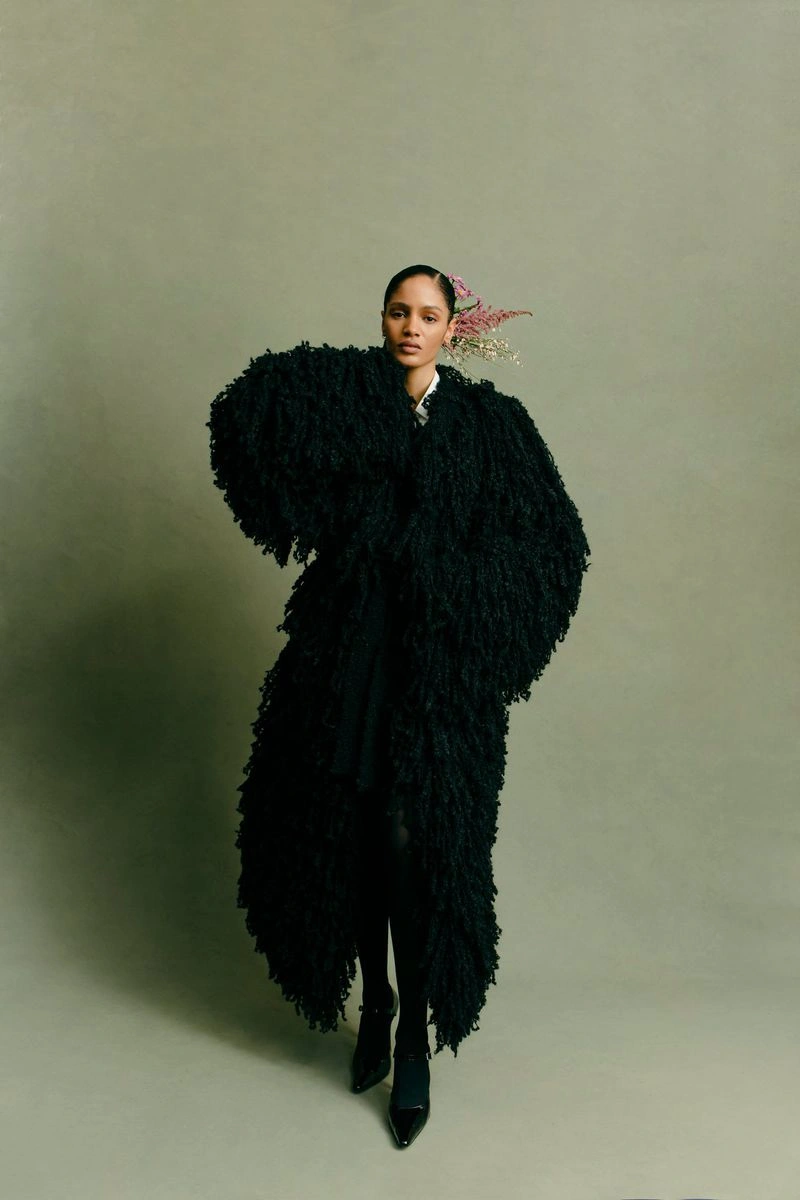 Model in textured black oversized coat wearing dark shoes against a muted olive backdrop