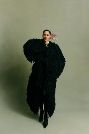 Model in textured black oversized coat wearing dark shoes against a muted olive backdrop