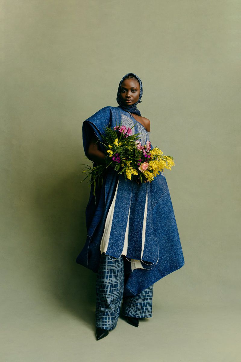 Model wearing blue draped wrap garment and patterned trousers holding a large floral bouquet against a muted grey-green backdrop.