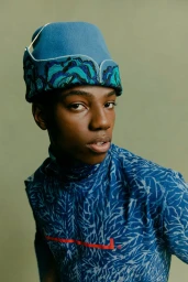 Close-up portrait of a Black male model wearing a blue patterned top and structured blue hat against a muted green backdrop.