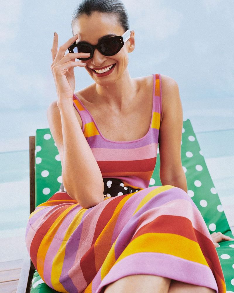 Smiling woman wearing a striped dress and sunglasses sitting on a beach chair with a polka dot pattern, with ocean in the background.