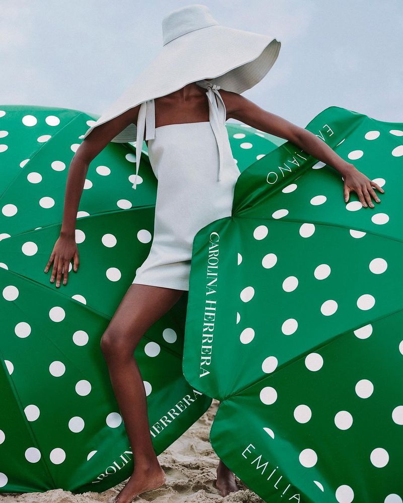Woman in white dress and sunhat on a sandy beach with green polka dot umbrellas, summer fashion editorial, Carolina Herrera branding.