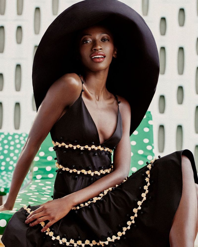 An African American woman poses in a black dress with white floral trim and a large black sun hat. The backdrop is a wall with oval designs.