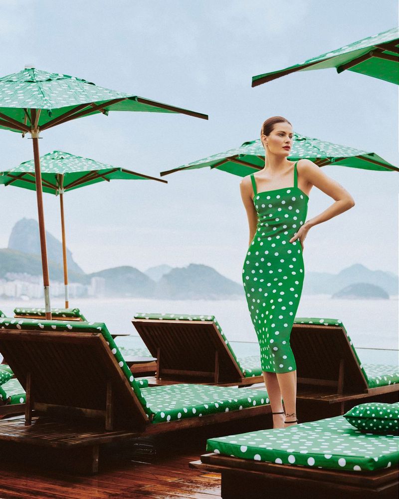 Woman in green polka dot dress poses on a deck chair with beach umbrellas, set against a coastal mountain backdrop, creating a fashion editorial.