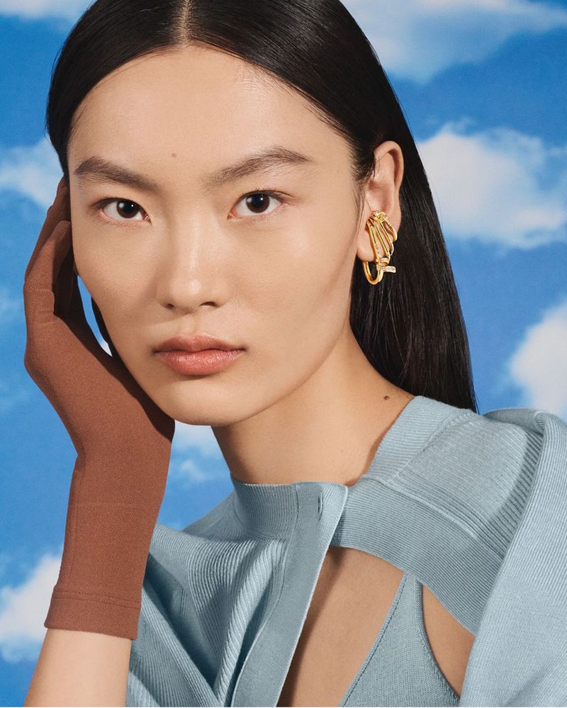 A woman poses wearing a light blue top, brown glove, and sculptural gold ear cuff against a sky-and-clouds background.
