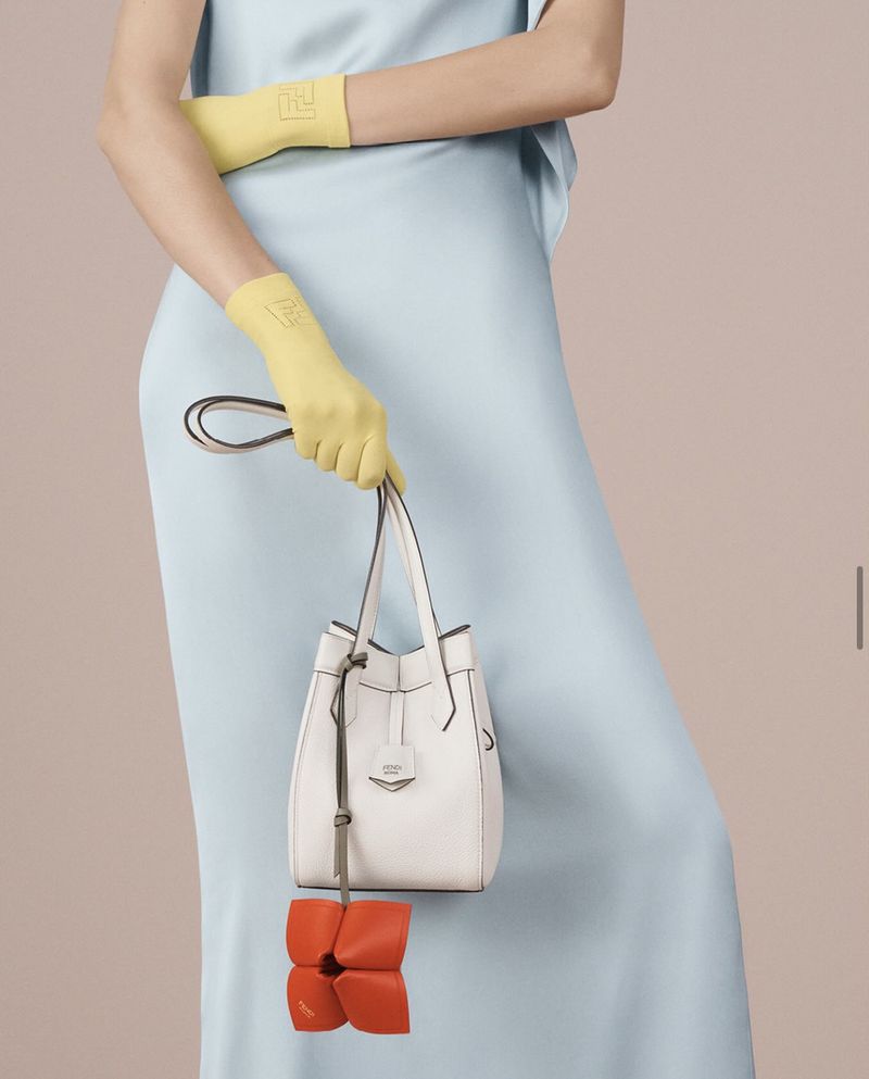 A person in a light blue dress holds a white leather purse with yellow gloves against a light tan background in a studio shot.